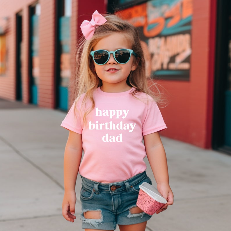 May include: A young girl wearing a pink t-shirt with the text "happy birthday dad" in white letters. She is wearing blue denim shorts and sunglasses.