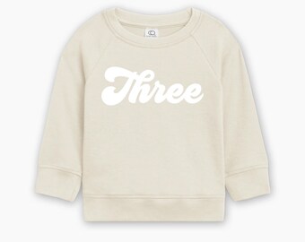 May include: Cream-colored toddler sweatshirt with the word "Three" in a white, retro font across the front. The long-sleeved top has a crew neck and ribbed cuffs and hem. The sweatshirt is made of a soft material.