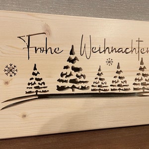Könnte beinhalten: Ein rechteckiges Holzschild mit den ausgeschnittenen Worten "Frohe Weihnachten". Das Schild zeigt eine Winterszene mit stilisierten Bäumen und Schneeflocken. Das Holz hat eine natürliche, helle Farbe.