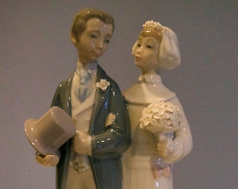 May include: A porcelain figurine of a bride and groom. The groom wears a dark teal jacket and holds a top hat. The bride wears a white dress with pink bow accents and a bouquet. The figurine is approximately 10 inches tall.