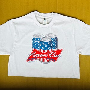 Puede incluir: Camiseta blanca con un gráfico de tres latas de cerveza envueltas en un diseño de bandera estadounidense. El texto "Ameri'Can" está impreso debajo de las latas.