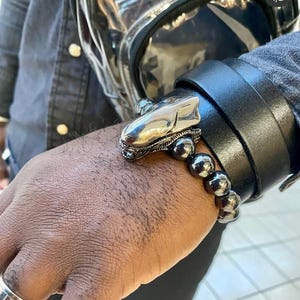 May include: A black leather bracelet with a silver alien head charm and a strand of dark gray beads. The bracelet is worn on a wrist, with a black jacket and a clear backpack in the background.