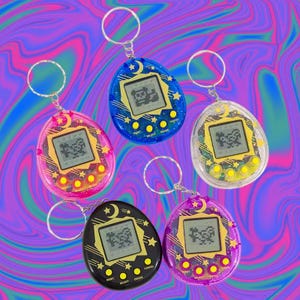 May include: Five colorful, retro handheld virtual pet game keychains. The keychains are in pink, blue, clear, black, and purple. Each has a small screen displaying a pixelated character, with buttons below. The keychains have a crescent moon and star design.