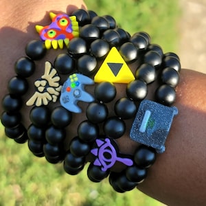 Pode incluir: Uma pilha de pulseiras feitas de contas pretas foscas com vários berloques coloridos, incluindo um triângulo amarelo, um controle de jogo azul, um símbolo de olho roxo, um emblema bege em forma de pássaro e uma máscara multicolorida com espinhos amarelos.