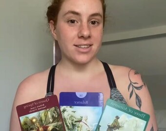 Puede incluir: Se muestran tres cartas del tarot, cada una con una ilustración y texto diferentes. Las cartas son de color rojo, azul y verde. La persona que sostiene las cartas lleva un sujetador deportivo negro.