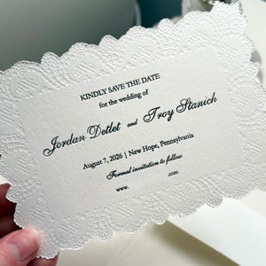 Könnte beinhalten: Eine weiße Hochzeitseinladung mit einem zarten, spitzenartigen Rand. Der Text enthält „Kindly Save the Date“ gefolgt von den Namen Jordan Dettet und Troy Stanich sowie dem Datum 7. August 2026 in New Hope, Pennsylvania.