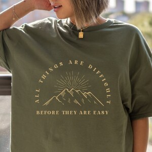 May include: A green t-shirt with a graphic of mountains and the text "All things are difficult before they are easy."