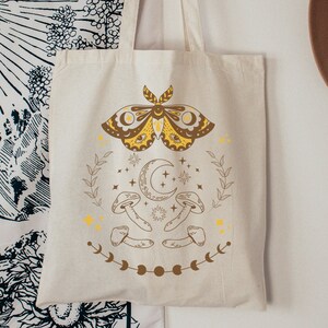 May include: A white canvas tote bag with a yellow and brown moth, crescent moon, and mushrooms graphic. The graphic is surrounded by stars and leaves.