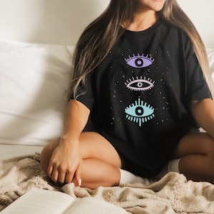 May include: A black t-shirt with a graphic of three stylized eyes in shades of blue, purple, and pink. The eyes are surrounded by small gold stars.