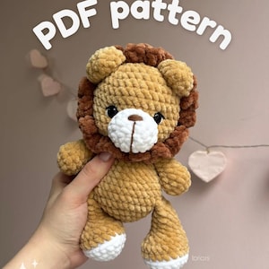 Può includere: Un leone all'uncinetto fatto a mano nei toni del beige e del marrone, con muso e zampe bianche. Il testo "PDF pattern" è visualizzato in un carattere giocoso. Il leone ha occhi neri e una criniera soffice. Il peluche è tenuto in una mano.