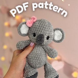 May include: A gray crocheted koala plush toy with a pink bow on its head, white inner ears, and dark gray feet. The text "PDF pattern" is displayed in white letters above the toy, which has black eyes and a dark gray nose.