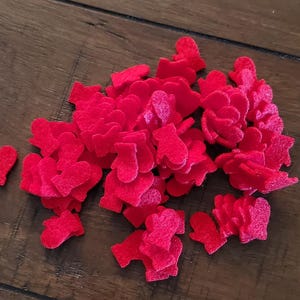 May include: A collection of small, bright red felt mittens scattered on a dark brown wooden surface. The mittens are uniformly shaped and appear to be craft supplies or decorative elements.