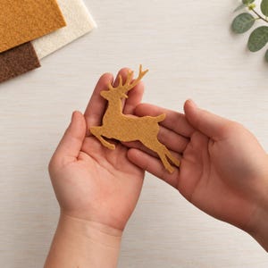 May include: A tan felt cutout in the shape of a leaping deer with antlers. The deer silhouette is crafted from a textured, non-woven fabric material and features a detailed profile with four legs extended in a mid-air jumping motion.