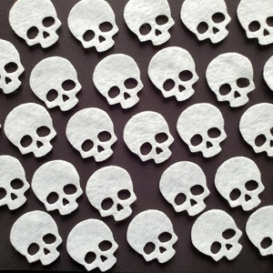 May include: A repeating pattern of white felt skull cutouts arranged in staggered rows against a dark brown background. Each skull shape features two circular eye sockets, a triangular nasal cavity, and a wide, toothless jaw opening.