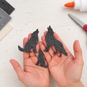 May include: Three dark gray felt cutouts in the shape of howling wolves. The felt shapes are textured and feature distinct silhouettes of wolves in a sitting or standing position, showcasing a craft material used for various decorative projects.