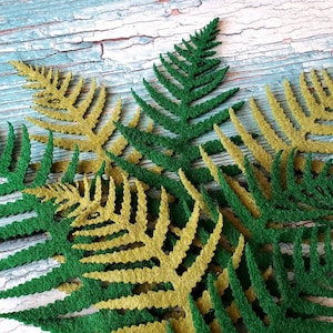 May include: A collection of flat, die-cut fern leaf shapes in alternating shades of forest green and metallic gold. The leaves feature intricate, serrated edges and a textured surface, arranged in an overlapping pattern to display their distinct colors.