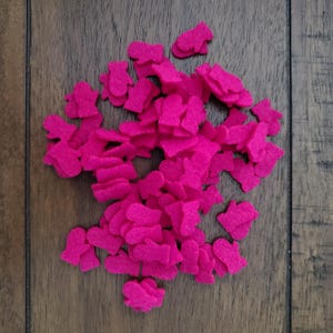 May include: A collection of small, hot pink felt shapes scattered on a wooden surface. The shapes appear to be cut-out figures, possibly for crafting or decorative purposes. The felt has a soft, matte texture, and the color is vibrant.