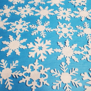 May include: A collection of white felt snowflakes scattered on a bright blue background. The snowflakes are various sizes and shapes, creating a festive winter scene.
