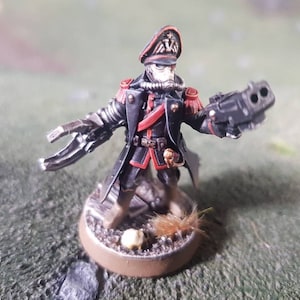 Puede incluir: Una figura en miniatura pintada de negro y rojo de un soldado que lleva un abrigo negro y un sombrero rojo y negro. La figura sostiene un arma negra con un cañón plateado. La figura está de pie sobre una base marrón con un pequeño parche de piel marrón.