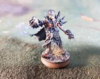 May include: A miniature figure of a warrior wearing black and silver armour with a skull helmet and a gas mask. The figure is holding a weapon in one hand and a chain in the other. The figure is standing on a brown base.