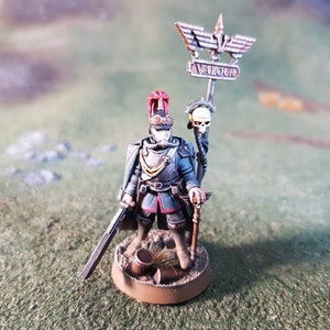 May include: A miniature painted figure of a soldier wearing a blue uniform and a red and gold helmet. The soldier is holding a sword and a staff with a skull on top. The staff also has a banner that reads "VALOUR".