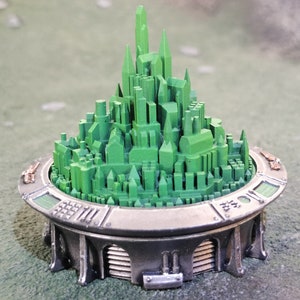 May include: A miniature model of a green city on a circular, metallic base with a detailed design. The city is made of small, rectangular buildings and towers.