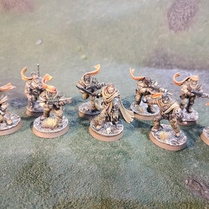 May include: A group of eight miniature figures painted in muted brown and tan colours, each wearing a hooded cloak with a bright orange scarf. The figures are posed in a variety of action stances, holding weapons and standing on round bases.