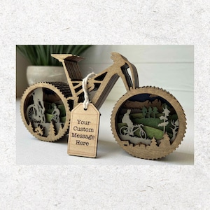 May include: A laser-cut wooden bicycle sculpture featuring layered mountain and forest silhouettes within the wheels. A small rectangular wooden tag hangs from the frame by a string, displaying the printed text: Your Custom Message Here.