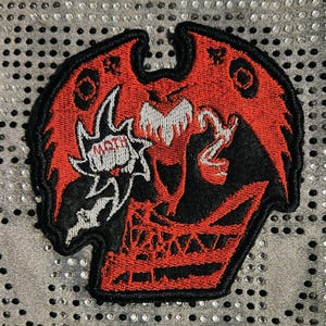 Può includere: Toppa ricamata con un disegno di falena rosso e nero. La toppa ha un bordo nero e la parola "MOTH" in bianco. Il disegno della falena ha un aspetto stilizzato, quasi demoniaco, con dettagli intricati.
