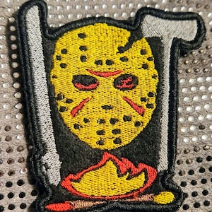 May include: Embroidered patch featuring a yellow hockey mask with red accents and black outlines. The patch includes a silver machete and a red flame design at the bottom. The patch is designed for horror movie fans.