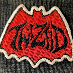 May include: A bat-shaped embroidered patch featuring a vibrant red background with a thick white border. The center displays the word TOZID stitched in a bold, stylized black font with sharp, angular edges that follow the bat wing contours.