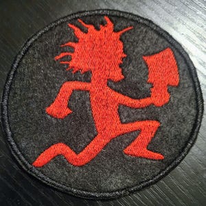 May include: A circular black patch featuring a red embroidered figure with an axe, in a running stance. The figure has spiky hair. The patch has a black border and is likely a logo or emblem.