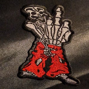Peut inclure: Un écusson brodé représentant une main squelettique faisant un geste du majeur. Le squelette porte un short rouge vif avec des détails noirs usés. Le motif est souligné par un fil noir, mettant en valeur des textures d'os blancs et gris.