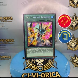 Puede incluir: Una carta de hechizo holográfica "The Gaze of Timaeus" del juego de cartas coleccionables Yu-Gi-Oh! La carta presenta una colorida ilustración de una criatura parecida a un dragón con texto en la parte superior e inferior. Se ven dos pegatinas circulares.