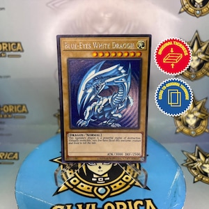 Op de afbeelding: Een Yu-Gi-Oh! ruilkaart met de Blue-Eyes White Dragon. De kaart heeft een blauwe en witte drakenillustratie met de tekst "BLUE-EYES WHITE DRAGON". De kaart wordt getoond op een blauwe standaard met een gouden ontwerp.