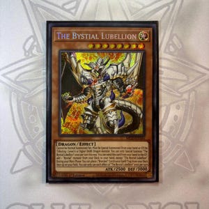 May include: A framed trading card featuring "The Bystial Lubellion" dragon. The card has a gold and brown background with a detailed illustration of a white and silver dragon. The card text includes "DRAGON/EFFECT" and other game instructions.