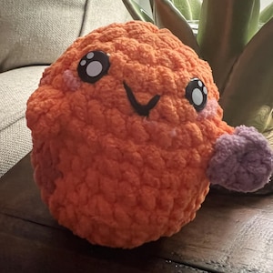 May include: A handmade orange crochet plushie with black and white eyes, pink cheeks, and a black stitched smile. A small purple crochet appendage extends from the side. The plushie is sitting on a dark wooden surface.