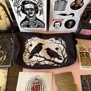 May include: A collection of gothic-themed merchandise, including a black zippered pouch with two crows perched on a branch, stickers, and cards. One sticker features Edgar Allan Poe. Other items include quote cards and tarot cards.