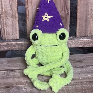 Può includere: Una rana di uncinetto verde che indossa un cappello da mago viola con una stella gialla. La rana è seduta su una superficie di legno.