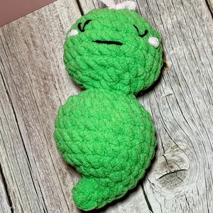 May include: A handmade, green crochet creature with a smiling face, white accents, and a small white bow. The plush toy is made of soft yarn and sits on a wooden surface. The toy has a rounded body and a small tail.