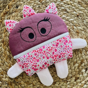 Könnte beinhalten: Ein Monster aus rosa-weißem Blumenstoff mit einer Reißverschlusstasche und zwei weißen, flauschigen Armen. Das Monster hat zwei große schwarze Augen mit Wimpern und einen burgunderfarbenen Stoffkörper.