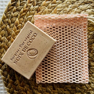 Peut inclure: Un sac en filet rose avec une fermeture à cordon et une perle en bois. Le sac contient une barre de savon rectangulaire avec le texte "Surgras - Pur végétal NOIX DE COCO" imprimé dessus.