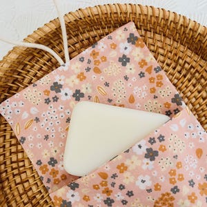 Pode incluir: Uma barra de sabão branca repousa em uma bolsa de tecido rosa floral, colocada dentro de uma cesta de vime. A bolsa apresenta um padrão de pequenas flores brancas, cinzas e laranja. Um cordão branco está preso.