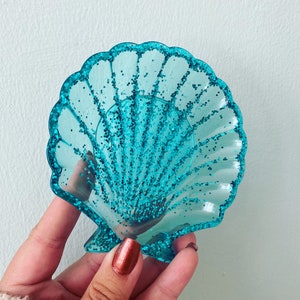 May include: A turquoise seashell-shaped trinket dish with glitter detailing. The dish is translucent and has a scalloped edge. The glitter adds a sparkling effect to the decorative piece. Ideal for holding small items, this dish is a charming accessory.