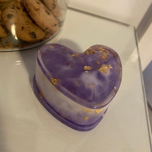 May include: A heart-shaped trinket box with a marbled purple and white design, accented with gold leaf. The box is on a white surface. A jar of chocolate chip cookies is in the background.