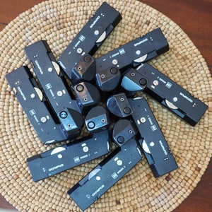 Puede incluir: Una colección de accesorios negros para cámaras Olympus, incluyendo bobinadoras y otros componentes. Los artículos están dispuestos sobre una estera circular tejida. Las bobinadoras están etiquetadas con "Winder 1" y "Winder 2" y el logotipo de Olympus.