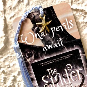 Peut inclure: Marque-page avec un pompon et un cordon bleu clair, et un charm en forme d'étoile de mer dorée. Le marque-page présente le texte "What perils await" et le titre "The Strider and the Regulus".