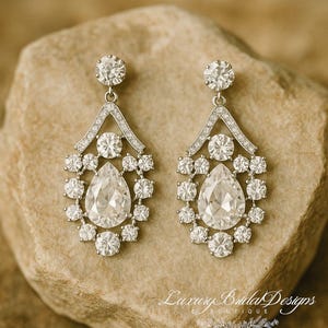 May include: A pair of silver-toned drop earrings featuring a central pear-shaped clear stone surrounded by a cluster of round clear stones, with a triangular top section encrusted with small clear stones. The text Luxury Bridal Designs is visible.