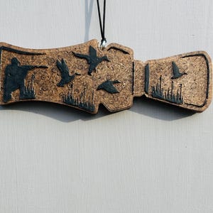 May include: A brown wooden ornament with a black cord for hanging. The ornament features a hunter aiming a gun at flying ducks, with additional ducks and marsh grass details. The design is carved into the wood.