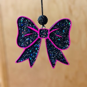 May include: A black glitter bow-shaped ornament with a bright pink outline. The bow is suspended by a black string and a small, black, beaded sphere. The glitter has flecks of blue, green, and silver.